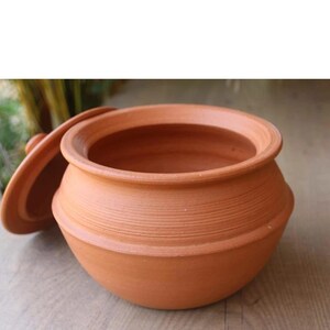 Puede incluir: Una olla de barro con tapa. La olla es redonda y tiene una abertura ancha. La tapa es ligeramente abovedada y tiene un pequeño pomo en la parte superior.