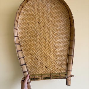 May include: A large, woven bamboo tray with a rounded top and a rectangular base. The tray features a herringbone weave pattern in natural tones. The frame is made of thicker bamboo strips, and the tray is held by a hand.