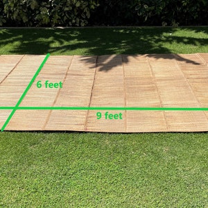 May include: A woven brown mat with a natural texture, measuring 6 feet by 9 feet. The mat is laid out on a green grassy lawn.