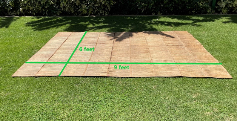 May include: A woven brown mat with a natural texture, measuring 6 feet by 9 feet. The mat is laid out on a green grassy lawn.