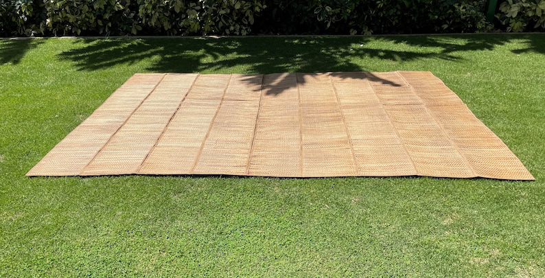 May include: A large, woven, natural-fiber mat, likely made of bamboo or straw, is laid out on a green grassy lawn. The mat is light brown in color and has a woven texture.
