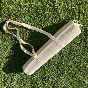 Pu&ograve; includere: Un porta-tappeto da yoga in tela bianca con un cinturino lungo. Il porta-tappeto &egrave; arrotolato e legato con uno spago in cima. Il porta-tappeto &egrave; sdraiato su un letto di erba verde.