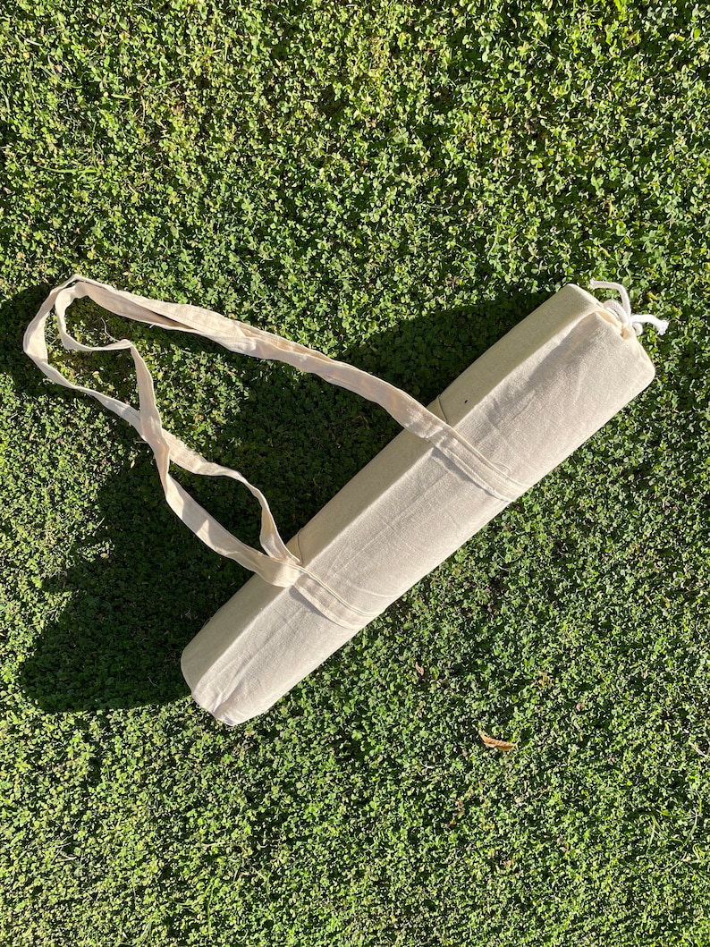 Pu&ograve; includere: Un porta-tappeto da yoga in tela bianca con un cinturino lungo. Il porta-tappeto &egrave; arrotolato e legato con uno spago in cima. Il porta-tappeto &egrave; sdraiato su un letto di erba verde.