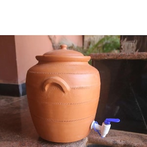 Może przedstawiać: Dystrybutor wody z terakoty z pokrywką i kranem. Okrągły, brązowy gliniany garnek ma zakrzywiony uchwyt i ozdobne szwy wokół korpusu. Kran jest biały z niebieskim uchwytem. Dystrybutor stoi na kamiennej powierzchni.