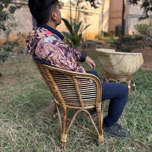 K&ouml;nnte beinhalten: Eine Person sitzt auf einem Korbstuhl mit einer Jacke mit Blumenmuster. Der Stuhl hat eine geflochtene R&uuml;ckenlehne und Armlehnen. Die Person tr&auml;gt eine schwarze Hose und schwarze Schuhe. Der Stuhl steht auf einer grasbewachsenen Fl&auml;che.
