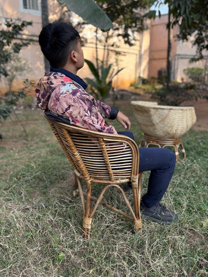 K&ouml;nnte beinhalten: Eine Person sitzt auf einem Korbstuhl mit einer Jacke mit Blumenmuster. Der Stuhl hat eine geflochtene R&uuml;ckenlehne und Armlehnen. Die Person tr&auml;gt eine schwarze Hose und schwarze Schuhe. Der Stuhl steht auf einer grasbewachsenen Fl&auml;che.