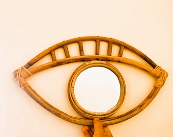 Könnte beinhalten: Ein Holzspiegel in Augenform mit einem runden Spiegel in der Mitte. Der Spiegel ist aus natürlichem geflochtenem Rattan gefertigt und hat eine hellbraune Farbe.