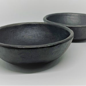 May include: Two black ceramic bowls, one slightly larger than the other, are shown on a white surface. The bowls have a smooth, slightly textured finish.
