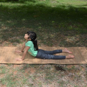 Pu&ograve; includere: Una persona giovane che pratica yoga su un tappetino in tessuto naturale all'aperto. Il tappetino &egrave; marrone e ha una trama naturale. La persona indossa una maglietta verde e leggings neri.