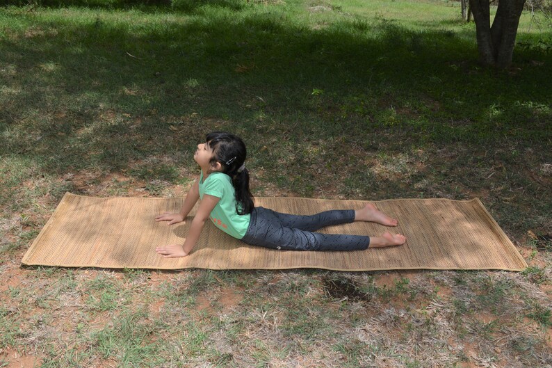 Pu&ograve; includere: Una persona giovane che pratica yoga su un tappetino in tessuto naturale all'aperto. Il tappetino &egrave; marrone e ha una trama naturale. La persona indossa una maglietta verde e leggings neri.
