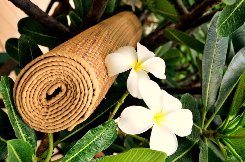 Pu&ograve; includere: Un tappetino da yoga marrone arrotolato con una trama intrecciata, appoggiato su foglie verdi e fiori bianchi. I fiori hanno cinque petali e un centro giallo.