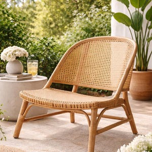 May include: A natural rattan chair with a woven seat and back. The chair has a curved back and armrests, and is set on a light-colored rug. A small side table with a vase of white flowers and a drink is visible.