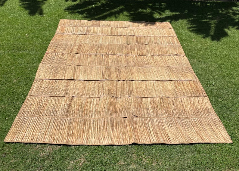 May include: A large, woven mat made of natural materials, likely straw or bamboo. The mat is light brown in color and has a rectangular shape. It is lying flat on a green grassy surface.
