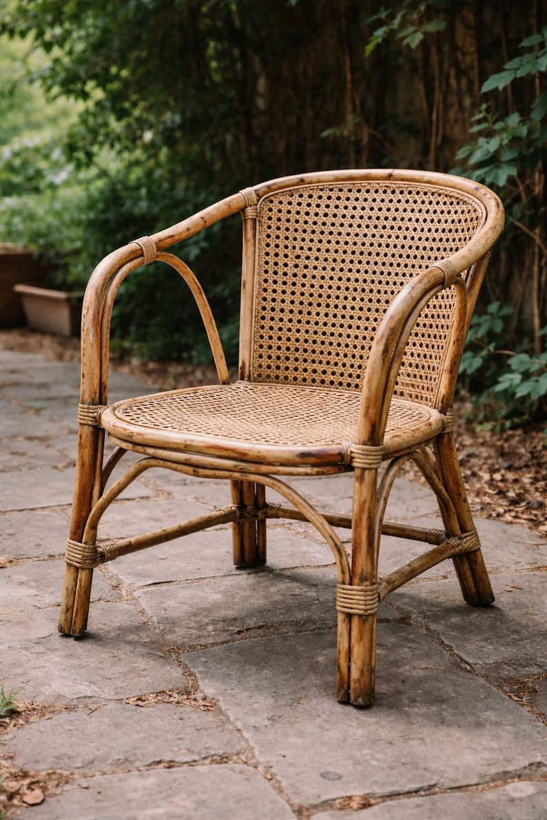 Op de afbeelding: Een natuurlijke rotan fauteuil met een geweven zitting en rugleuning. De stoel heeft gebogen armleuningen en poten en is geconstrueerd met geweven rotan binding. De stoel staat buiten op een stenen terras, met groen op de achtergrond.