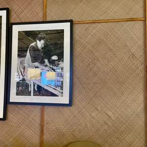 May include: A black framed photograph of a person in a white shirt and apron working at a table with a variety of items. The photograph is hanging on a wall with a woven texture.