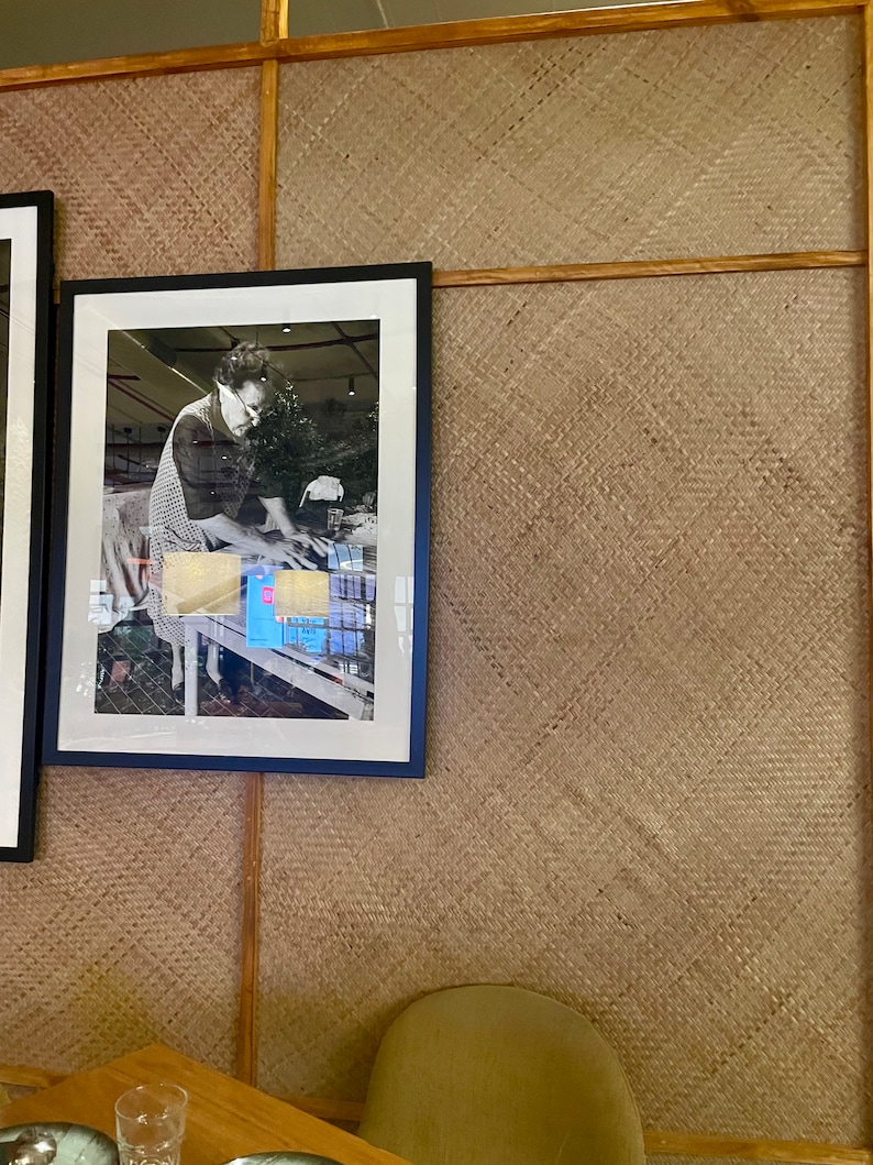 May include: A black framed photograph of a person in a white shirt and apron working at a table with a variety of items. The photograph is hanging on a wall with a woven texture.