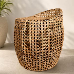 May include: A barrel-shaped stool crafted from light brown woven rattan with a natural finish. The piece features an open octagonal cane webbing pattern throughout its cylindrical body and a solid, slightly curved top surface for seating.