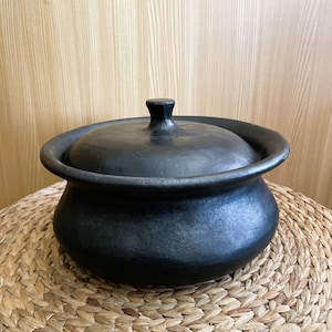 May include: A black ceramic cooking pot with a matching lid. The pot has a rounded body and a wide rim. The lid has a small, square handle. The pot is sitting on a woven, circular mat.