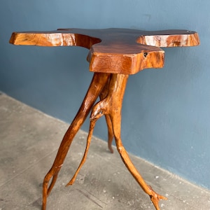 May include: A wooden table with a unique, natural, tree root base. The table top is made of a single piece of wood with a natural edge and a brown finish.