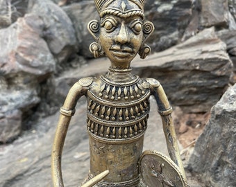 May include: A brass figurine of a seated figure wearing a headdress and holding a small drum. The figure is intricately detailed with a patterned chest piece and a detailed face.