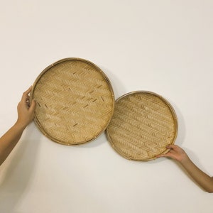 Pode incluir: Duas bandejas redondas de bambu trançado, uma maior que a outra, são seguradas contra uma parede branca.