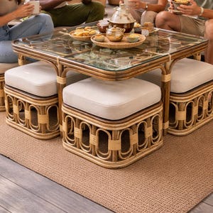 May include: A square coffee table with a glass top and a brown rattan frame, featuring four matching square ottomans with off-white upholstered cushions that tuck neatly underneath the table frame when not in use.