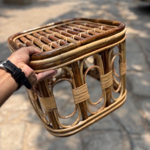 May include: A rectangular stool constructed from dark brown and light tan natural rattan. The seat features a slatted design, while the base is composed of arched supports reinforced with tightly wound rattan bindings at the joints.