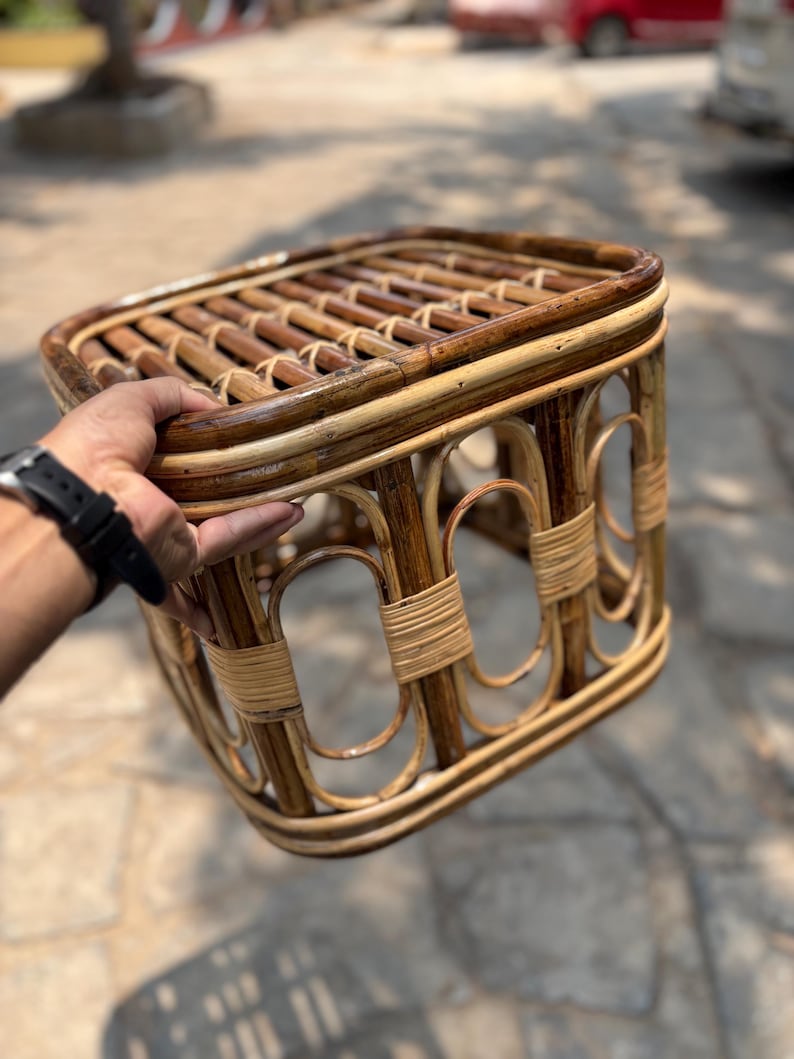 May include: A rectangular stool constructed from dark brown and light tan natural rattan. The seat features a slatted design, while the base is composed of arched supports reinforced with tightly wound rattan bindings at the joints.