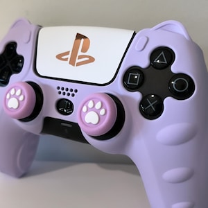 May include: A lavender purple PlayStation 5 controller with paw print button caps. The controller has a white faceplate with a gold PlayStation logo.