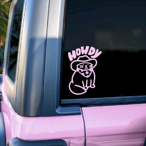 Pu&ograve; includere: Un adesivo in vinile rosa con un gatto dei cartoni animati che indossa un cappello da cowboy e la parola "HOWDY" sopra. L'adesivo &egrave; sul lunotto posteriore di un'auto rosa.