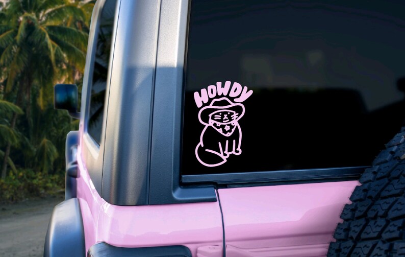 Pu&ograve; includere: Un adesivo in vinile rosa con un gatto dei cartoni animati che indossa un cappello da cowboy e la parola "HOWDY" sopra. L'adesivo &egrave; sul lunotto posteriore di un'auto rosa.
