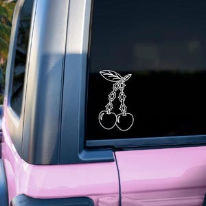 May include: White vinyl decal of two cherries with stems and leaves, applied to the rear window of a pink vehicle. The design is a simple outline, suitable for car decoration. The background shows a tropical landscape.