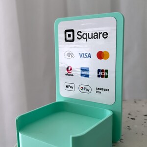 May include: A teal blue Square stand with a white sign that says "Square" and displays logos for various payment methods including Visa, Mastercard, American Express, Discover, JCB, Apple Pay, Google Pay, and Samsung Pay.