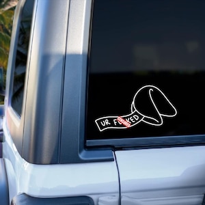 May include: A white vinyl decal applied to a vehicle window featuring a line art illustration of a fortune cookie with a paper strip extending from it. The text on the paper reads UR F*CKED with a red censored bar over the letter U.
