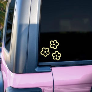 May include: Three yellow flower stickers are on the rear window of a pink Jeep. The stickers are simple, daisy-like designs. The vehicle is parked outdoors with a tropical background.