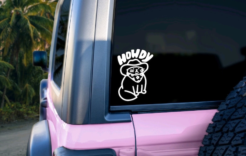 Pu&ograve; includere: Un adesivo in vinile bianco di un gatto che indossa un cappello da cowboy e un bandana con la parola "HOWDY" sopra. L'adesivo &egrave; sul lunotto posteriore di un'auto rosa.