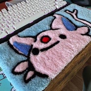 May include: A rectangular tufted rug featuring a stylised pink creature with blue ears, a red circular eye, and black outlines. The background is light blue, and the texture is thick and plush, designed to sit in front of a computer keyboard.