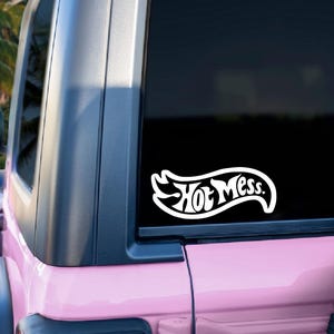 May include: White vinyl sticker with the words "Hot Mess" in a stylized font, applied to the rear window of a pink vehicle. The sticker is in a banner shape. Palm trees are visible in the background.