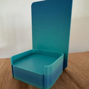 May include: A teal blue 3D printed phone stand with a square base and a tall rectangular back.