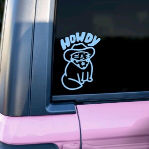 Pu&ograve; includere: Un adesivo in vinile bianco con un gatto dei cartoni animati che indossa un cappello da cowboy e un bandana. L'adesivo dice "Howdy" sopra il gatto.