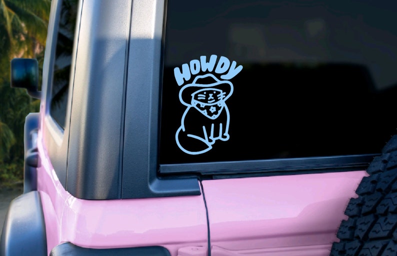 Pu&ograve; includere: Un adesivo in vinile bianco con un gatto dei cartoni animati che indossa un cappello da cowboy e un bandana. L'adesivo dice "Howdy" sopra il gatto.