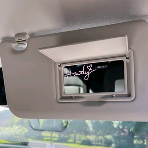 May include: A white car sun visor with a small mirror. The mirror has a pink sticker with the word "Howdy" and a heart.