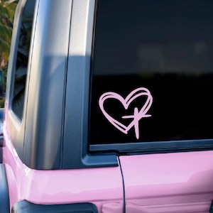 May include: A pink heart and cross sticker is displayed on the rear window of a pink vehicle. The sticker shows a hand-drawn style heart shape with a cross inside. The vehicle is parked outside with palm trees visible in the background.