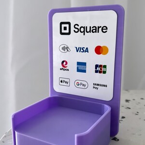 May include: A purple Square stand with a white sign that says "Square" and displays logos for Visa, Mastercard, American Express, Discover, JCB, Apple Pay, Google Pay, and Samsung Pay.