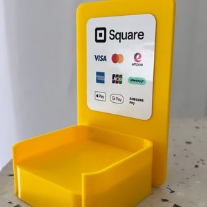 May include: A yellow plastic stand with a Square logo and a list of accepted payment methods including Visa, Mastercard, American Express, Discover, JCB, Afterpay, Apple Pay, Google Pay, and Samsung Pay.