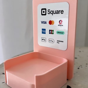 May include: A pink Square stand with a white label that says "Square" and lists accepted payment methods including Visa, Mastercard, American Express, Discover, JCB, eftpos, Afterpay, Apple Pay, Google Pay, and Samsung Pay.