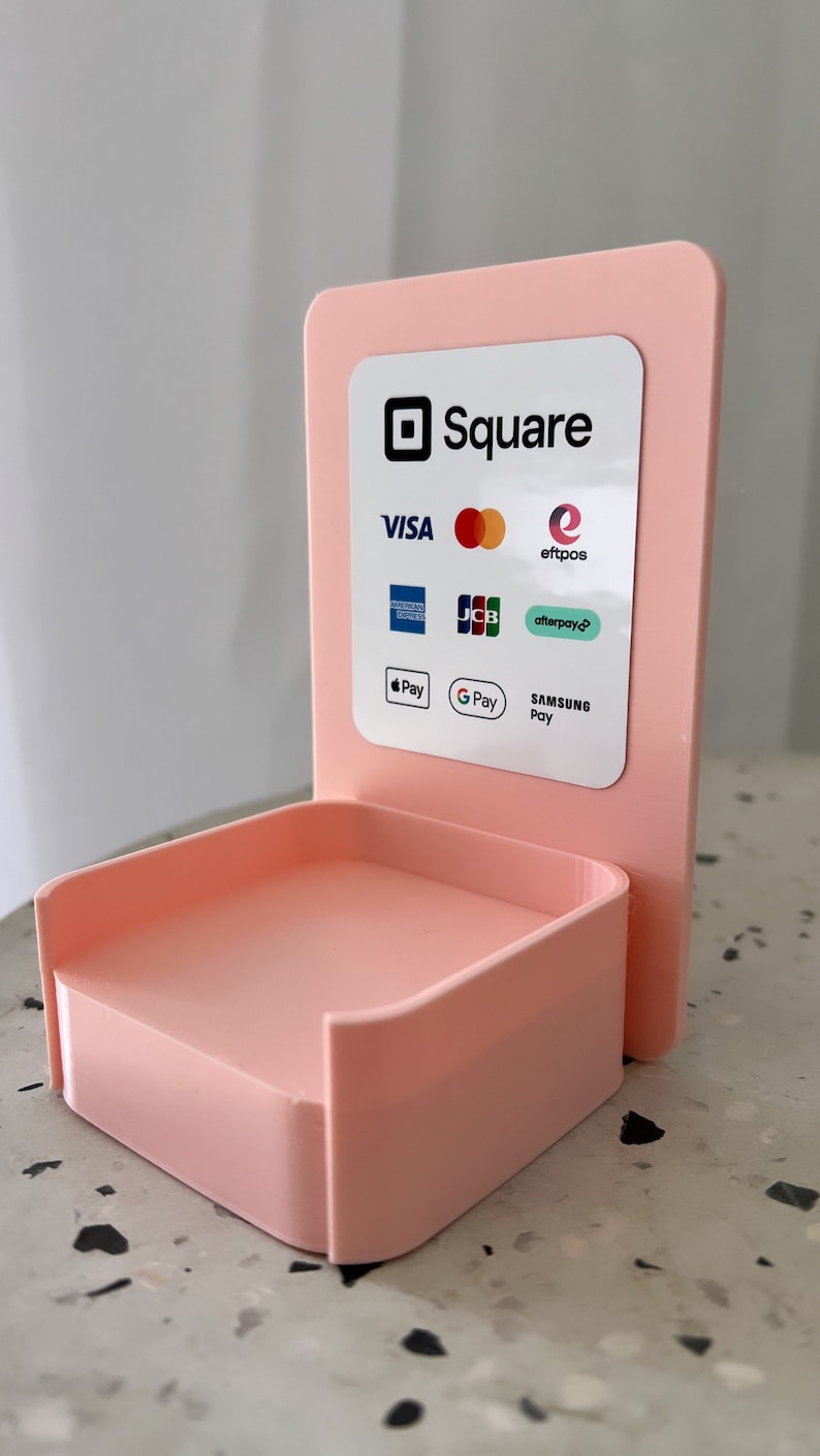 May include: A pink Square stand with a white label that says "Square" and lists accepted payment methods including Visa, Mastercard, American Express, Discover, JCB, eftpos, Afterpay, Apple Pay, Google Pay, and Samsung Pay.