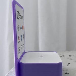 May include: A purple Square payment terminal stand with a white top and a side panel displaying the Square logo and accepted payment methods, including Visa, Mastercard, Discover, American Express, and Apple Pay.