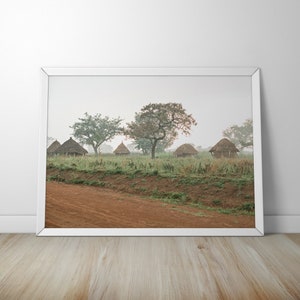 May include: A misty landscape with a row of thatched huts in a field.  The huts are made of straw and have a conical shape.  There are trees in the background and a dirt road in the foreground.