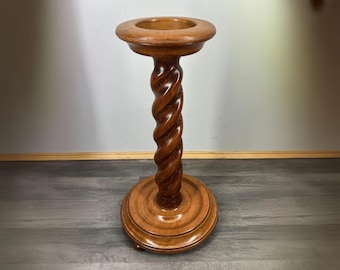 May include: A wooden plant stand with a carved, twisted column. The stand has a round base and a circular top with a recessed area. The wood has a warm, medium brown tone, and the stand appears to be vintage or antique.